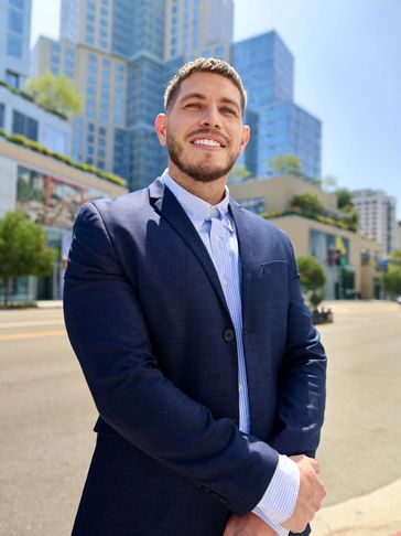 Confident man in a navy blazer smiling on a city street with tall buildings behind him.