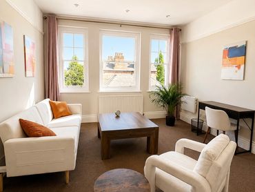 Bright living room with white furniture, wooden tables, and large windows letting in natural light.