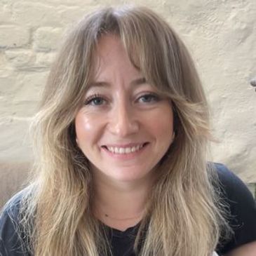 Smiling woman with blonde hair sitting indoors against a light brick wall.