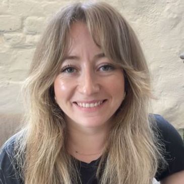 Smiling woman with blonde hair sitting indoors against a light brick wall.