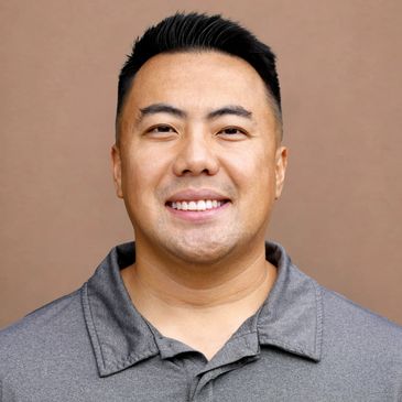 Smiling man in a gray collared shirt against a plain brown background.