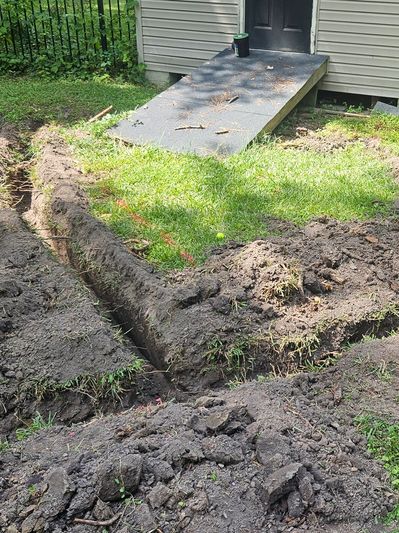 Freshly dug trench near a house ramp in a green yard.