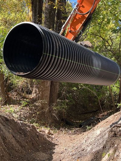 Large black corrugated pipe suspended by construction machinery over a dirt trench in a wooded area.