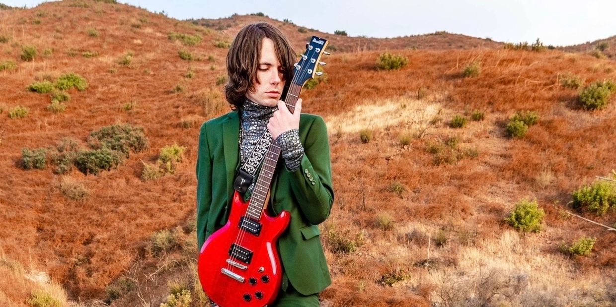 Person in green suit holding red electric guitar in dry hilly landscape.