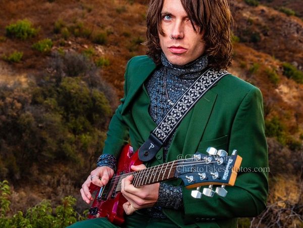 Musician in a green suit playing red electric guitar outdoors.
