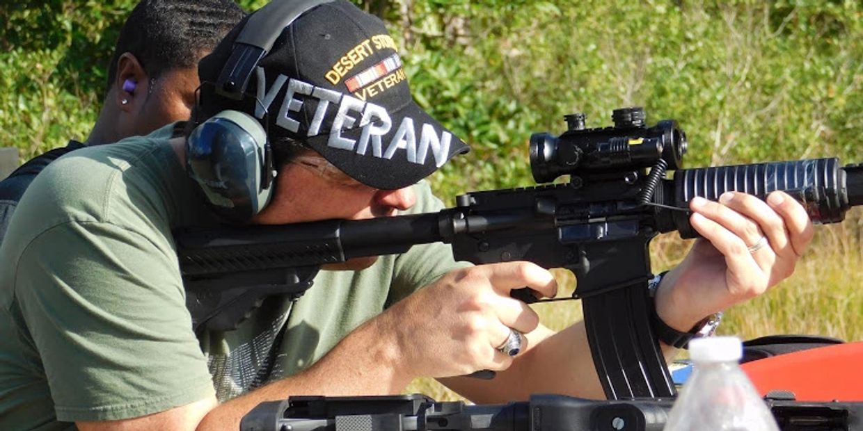 Veteran aiming a rifle at an outdoor shooting range, wearing ear protection and a 'Desert Storm Veteran' hat.
