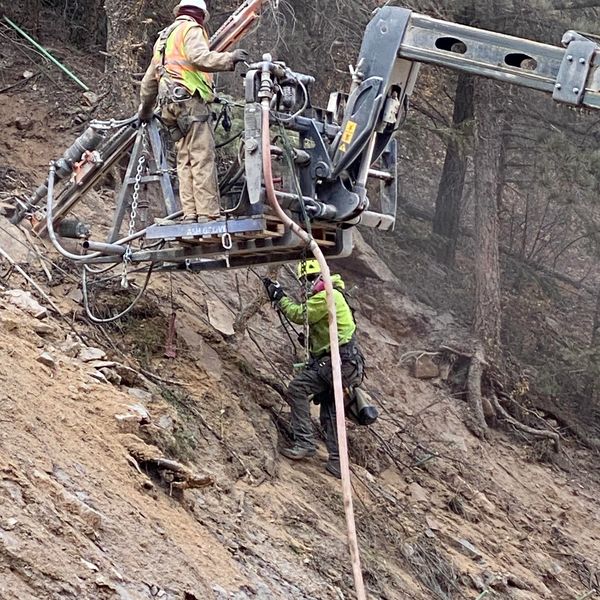 Uinta crew drilling rockfall anchors from a crane-held drill basket on a steep slope