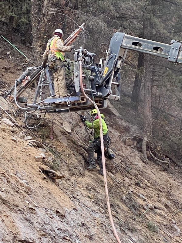 Two workers operating machinery on a steep, rocky hillside in a forest.