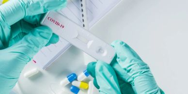 Hands with gloves holding a COVID-19 test with pills and notebook on table.