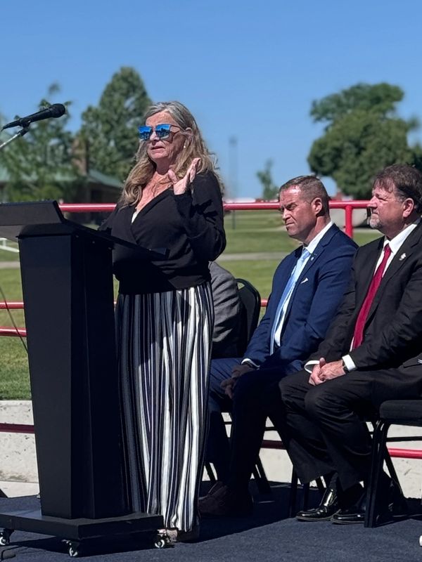 Woman in sunglasses speaks at podium while two men in suits listen.
