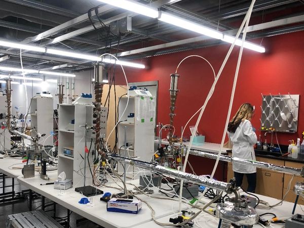 A woman in a lab coat works in a complex laboratory filled with scientific equipment and tubing.