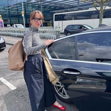 Woman in striped top and glasses stands by a black car, smiling.