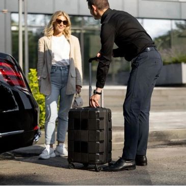 Man helps woman with luggage near a car outside a building.