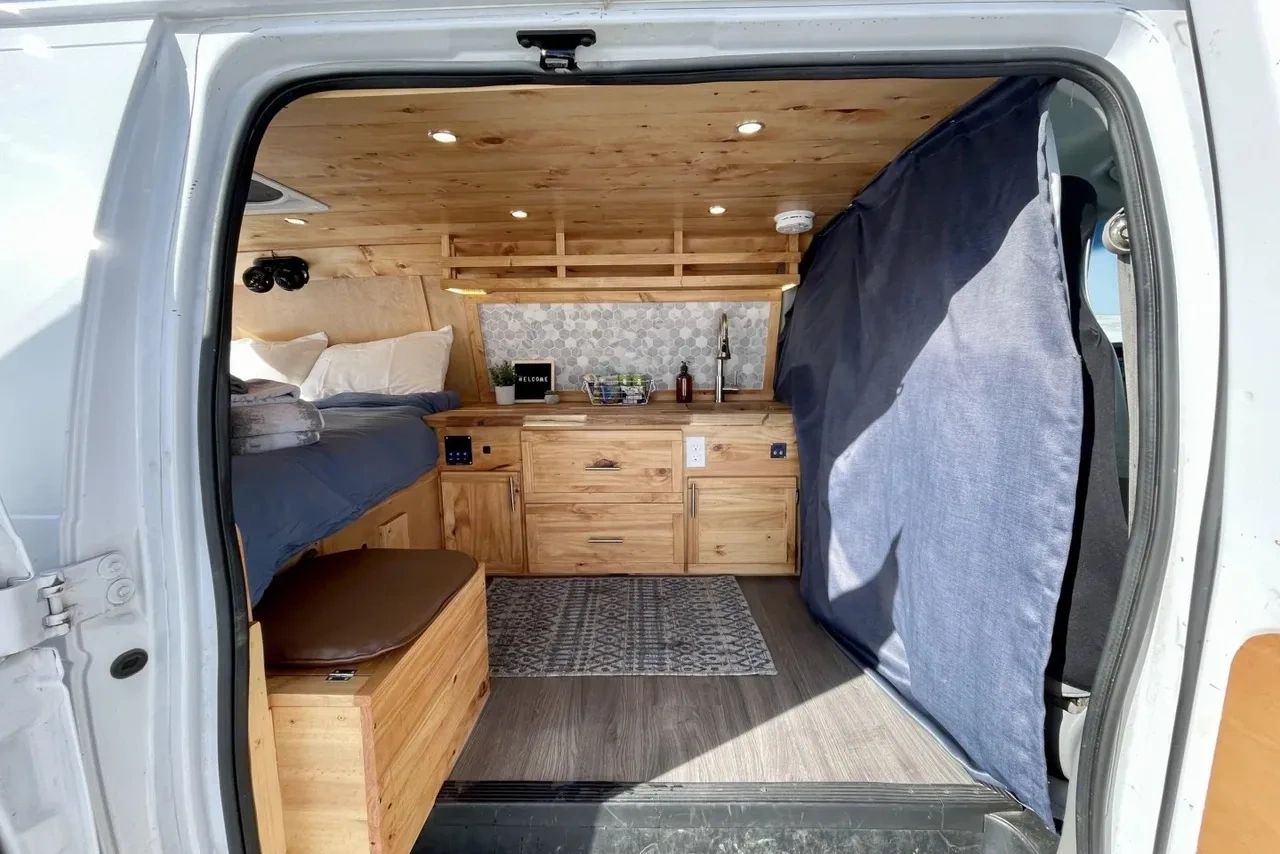 Cozy wooden camper van interior with a bed, kitchen, and seating area.