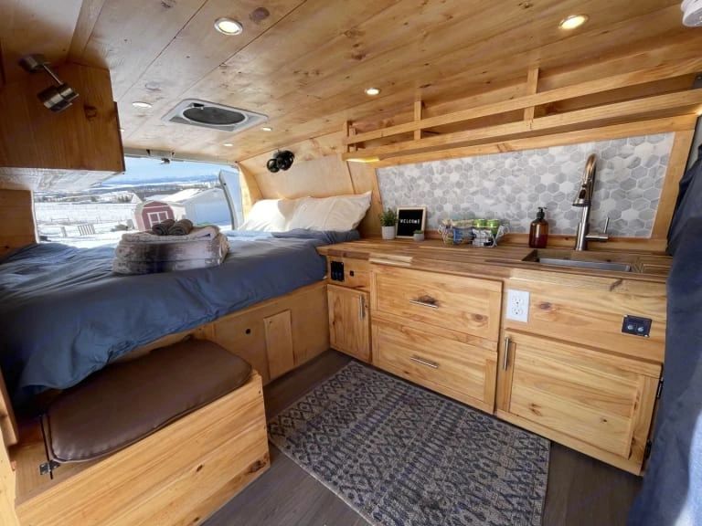Cozy wooden campervan interior with bed, kitchenette, and scenic window view.