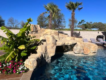 A sunny backyard pool with a rock waterfall and tropical plants.