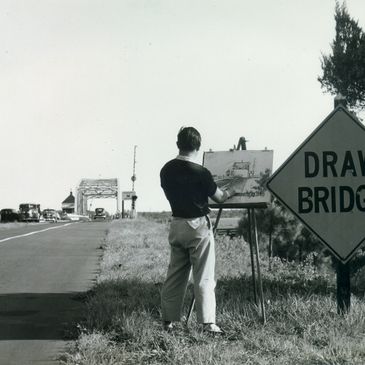 Man painting a drawbridge scene outdoors near a road sign.