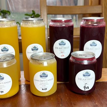 Bottles and jars of colorful juice labeled Unicorn Candy and Heartbeet on a wooden table.