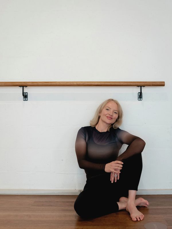 Woman in black sitting on wooden floor against white wall with ballet barre.