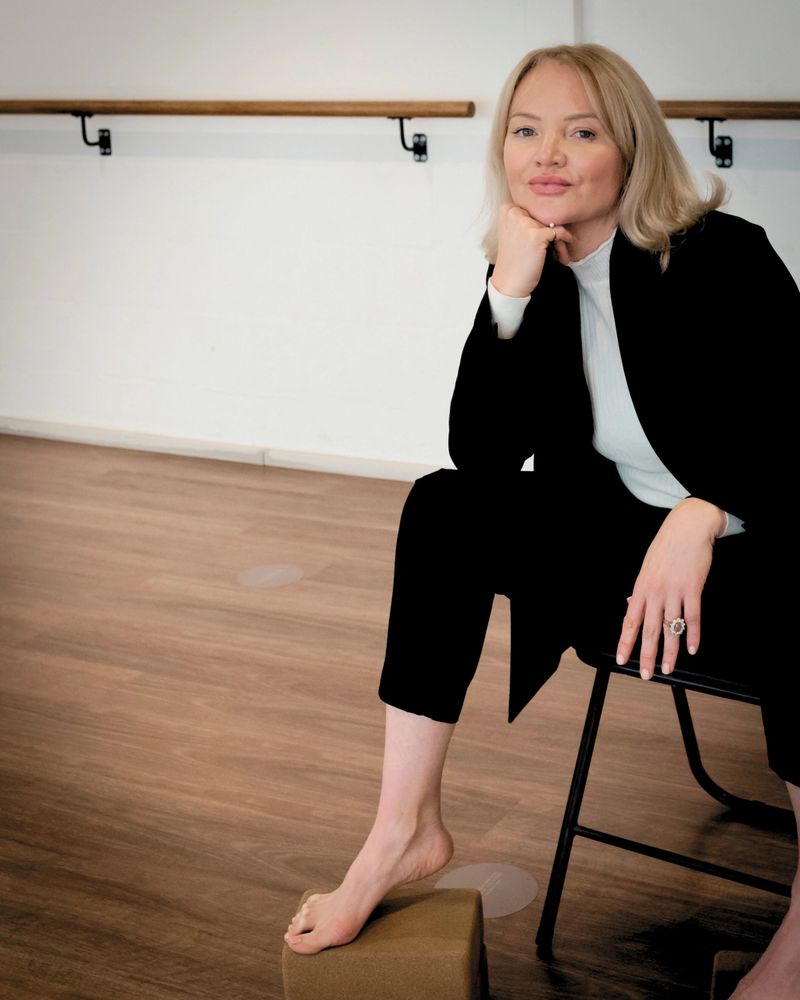Woman in black suit sitting barefoot on a chair in a dance studio.