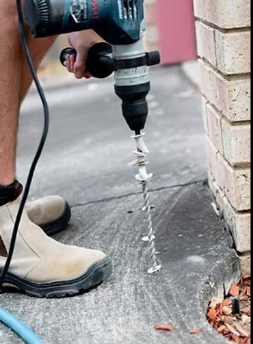 Person using a drill with an auger bit on concrete near a brick wall.