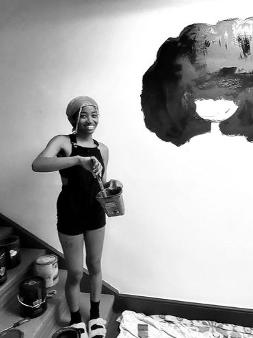 A young girl with a paintbrush  poses beside the mural she had just begun