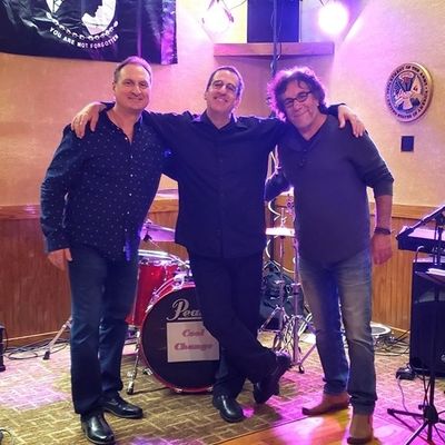 Three men posing happily in front of a drum set on stage.