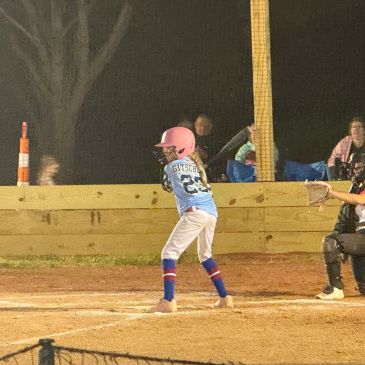 Young girl in pink helmet batting in a night softball game.