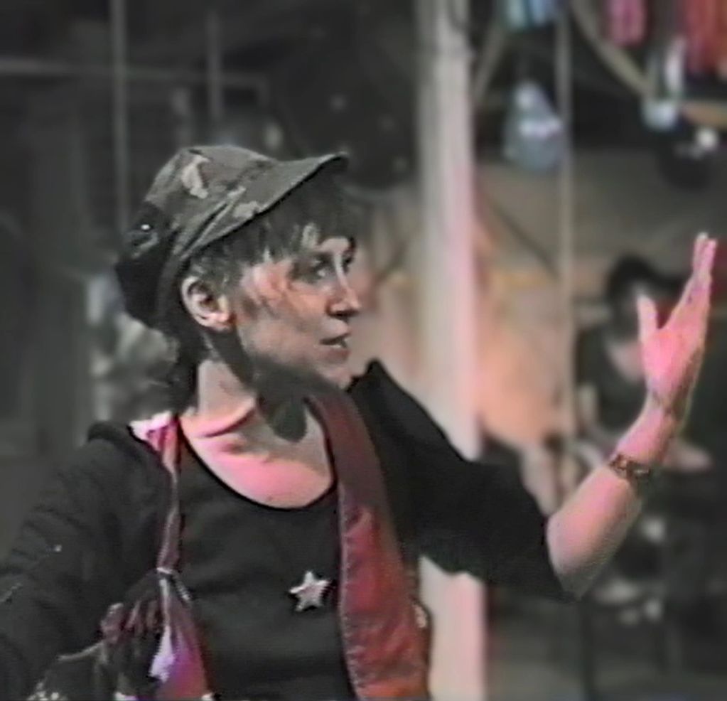 Person gesturing with one hand, wearing a cap and a star on their shirt.