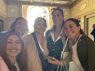 Five women smiling together indoors under ornate architecture.