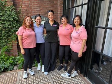 Five women standing together, smiling, dressed in casual attire with three wearing pink shirts.