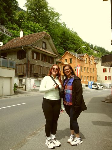 Two women standing on a street with houses and greenery in the background.