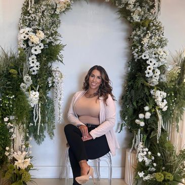 Woman sitting on a clear chair under a lush floral arch smiling.