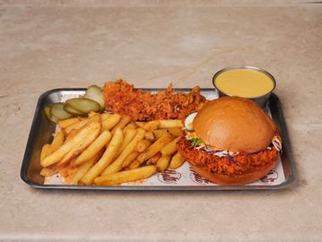 Spicy fried chicken sandwich with fries, pickles, and dipping sauce on a metal tray.