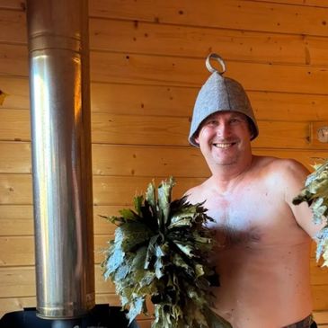 Man smiling in a sauna holding leafy branches, wearing a traditional sauna hat.