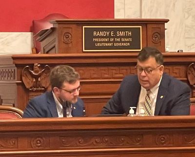 Two men in suits converse at a government chamber with a nameplate for Randy E. Smith.