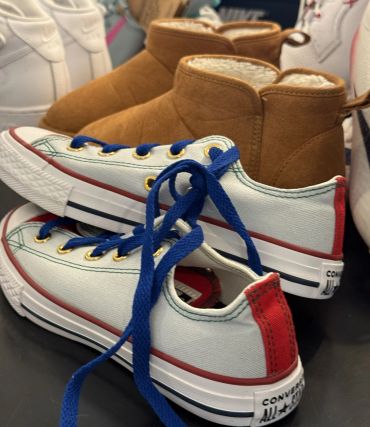 Pair of white Converse sneakers with blue laces and brown boots in the background.