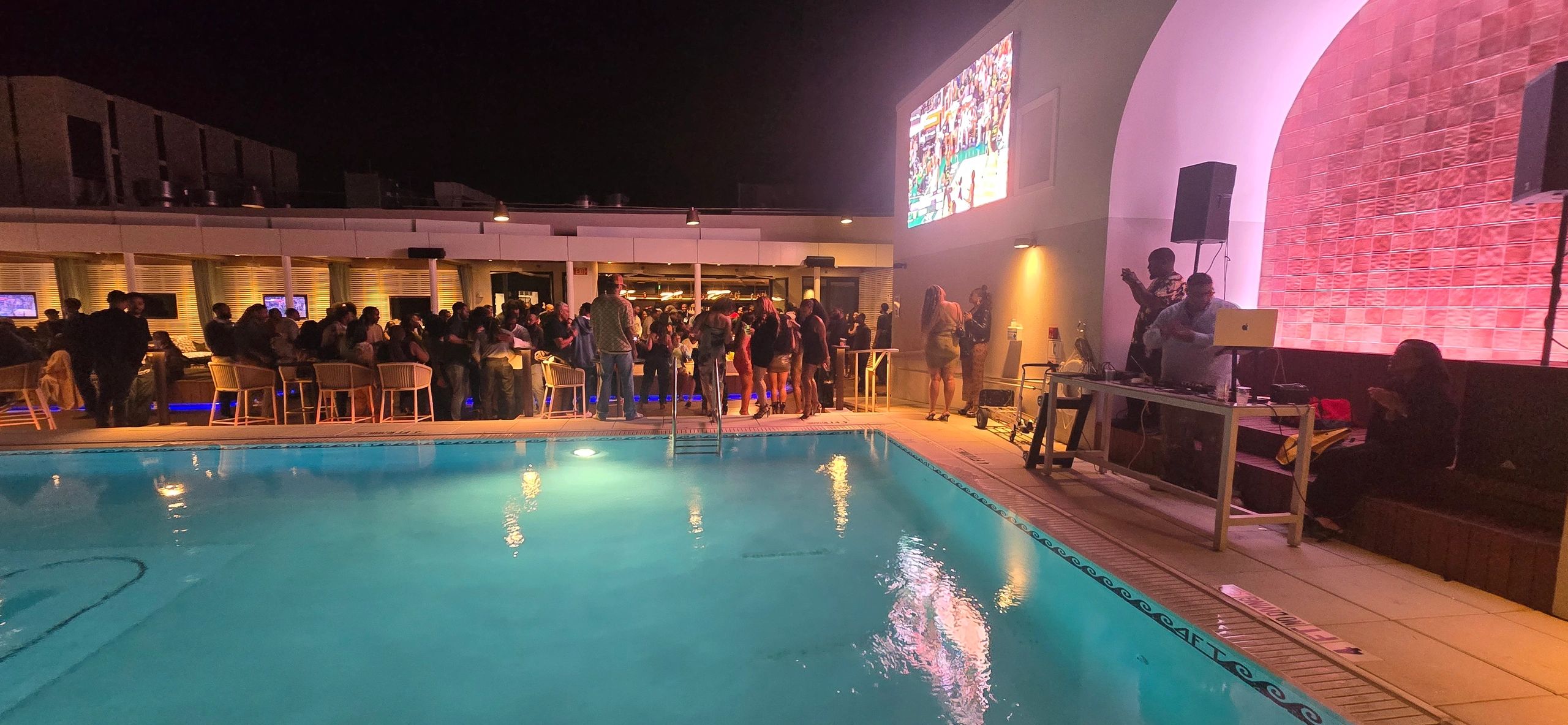Night pool party with crowd, DJ, and sports on large screen.