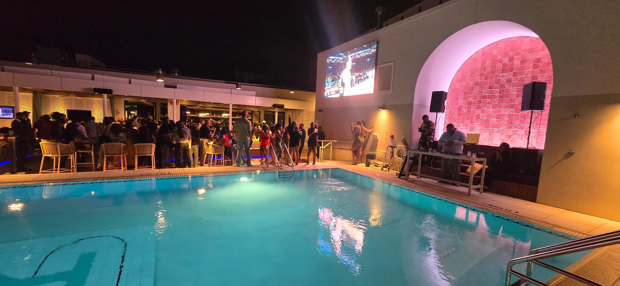 Night pool party with crowd, DJ, and large screen.