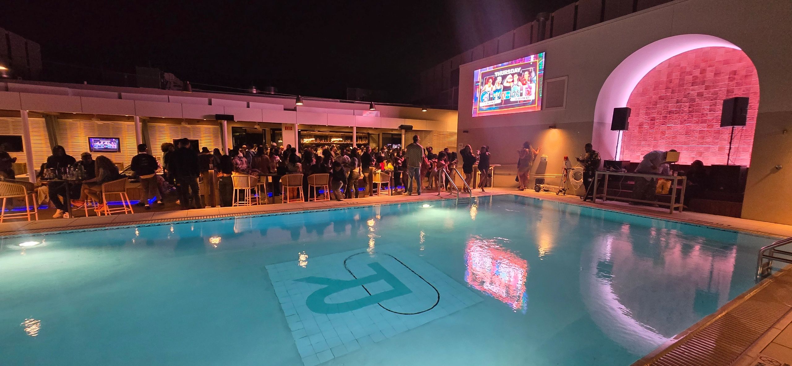 Night party by a pool with people socializing and colorful lights.