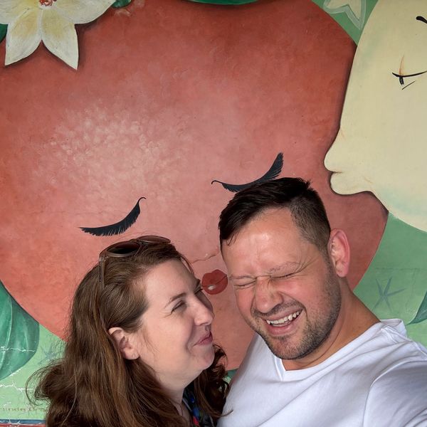 Couple smiling playfully in front of a colorful mural with faces.
