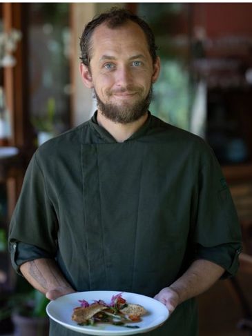 Homem sorridente com jaleco preto segurando um prato de comida.