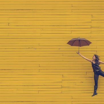 A joyful woman jumping with an umbrella against a yellow wooden wall.