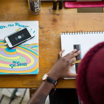 Person measuring a sketchbook page with a ruler at a wooden desk.
