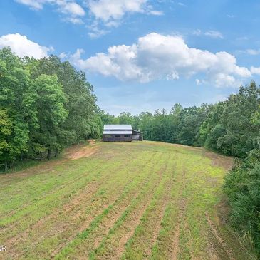 A small barn at the end of a grassy field surrounded by trees under a blue sky.