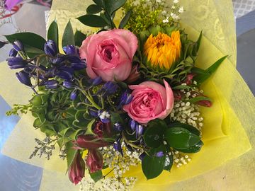 A vibrant bouquet with pink roses, yellow and purple flowers wrapped in yellow paper.