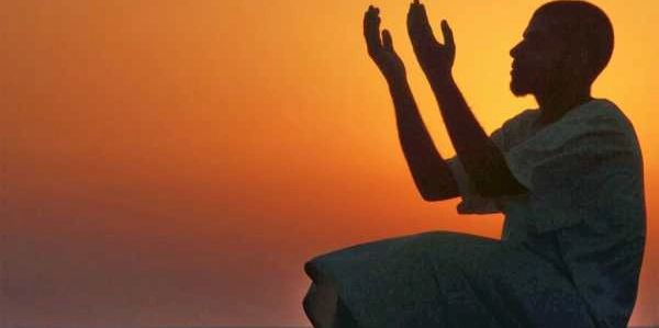 Silhouette of person praying at sunset with hands raised.