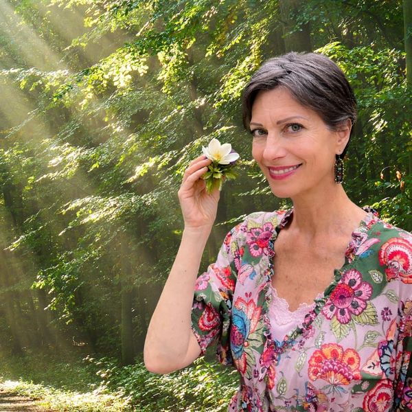 A woman in a floral dress holding a white flower in a sunlit forest.