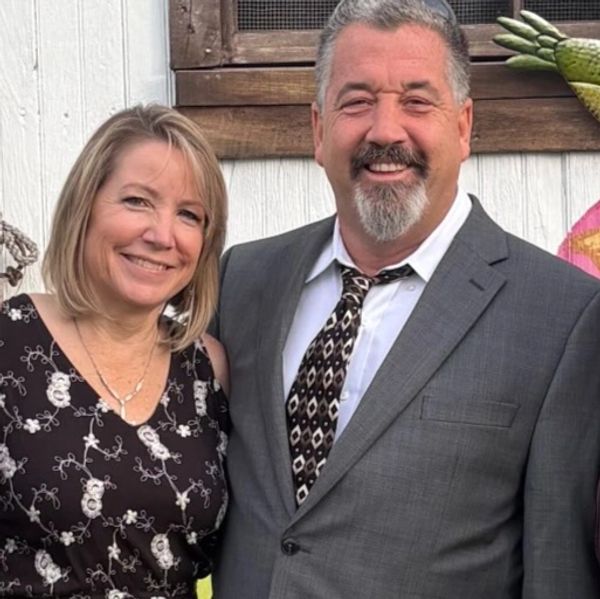 Smiling couple dressed formally, standing outdoors by a white wall.