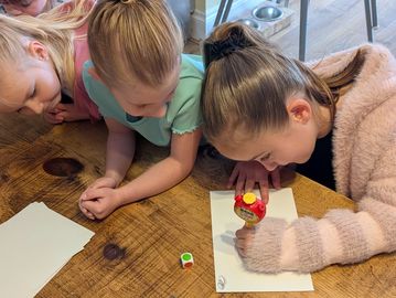 Three children leaning on a table, playing with a toy and watching closely.